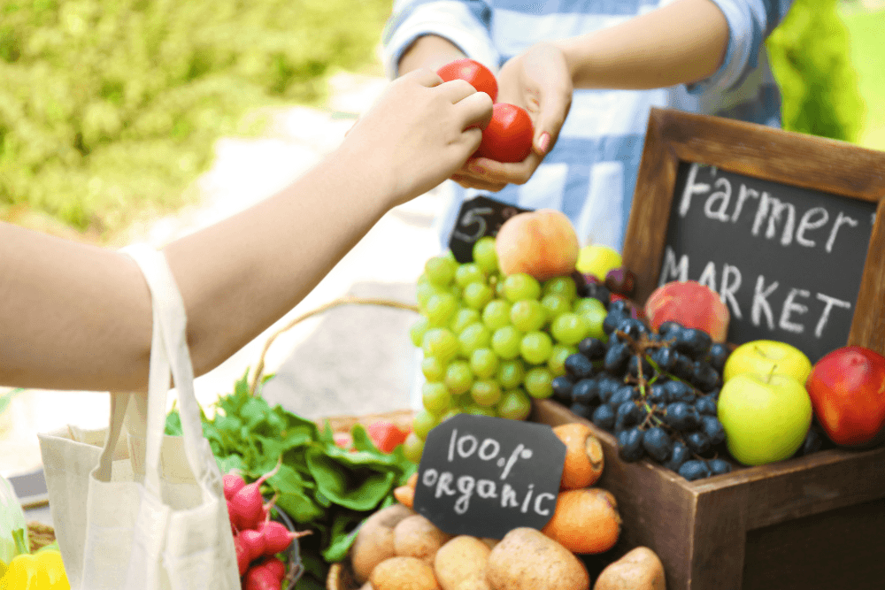 A farm market table full of fruits and vegetables and a woman handing two tomatoes to another woman