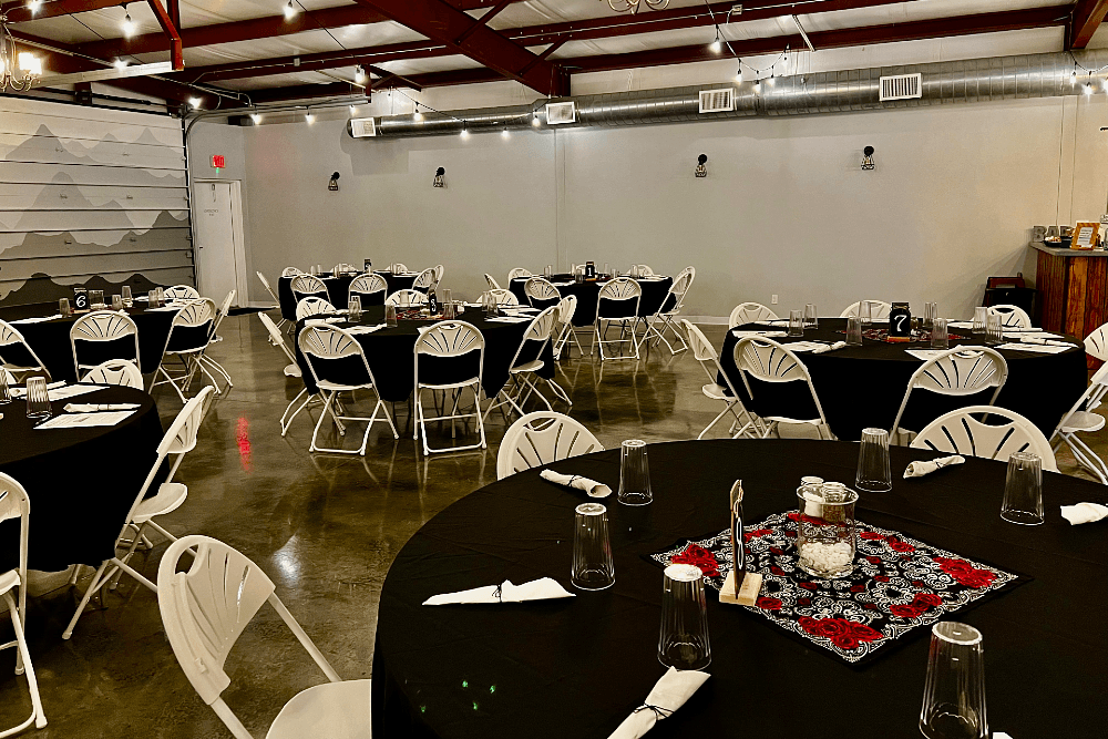 A large hall space with two garage doors, string lights and round wood tables with white folding chairs