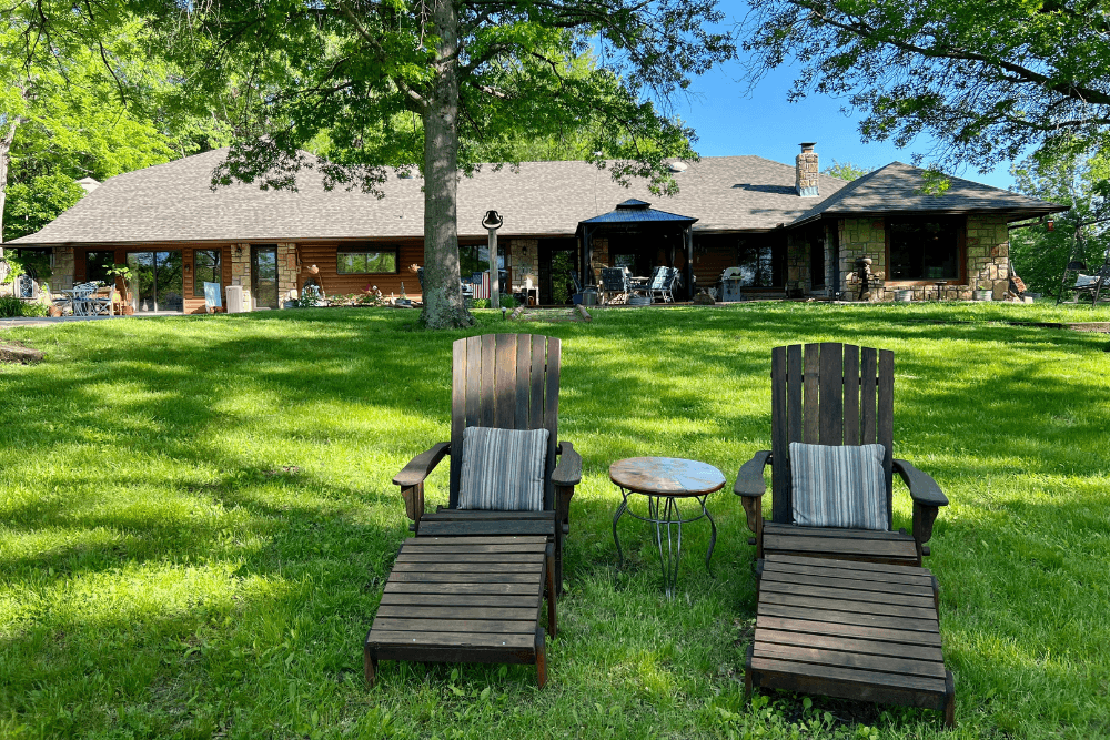 Large home with stone detail, brown roof, expansive lawn with gazebo, patio furniture and two adirondack chairs