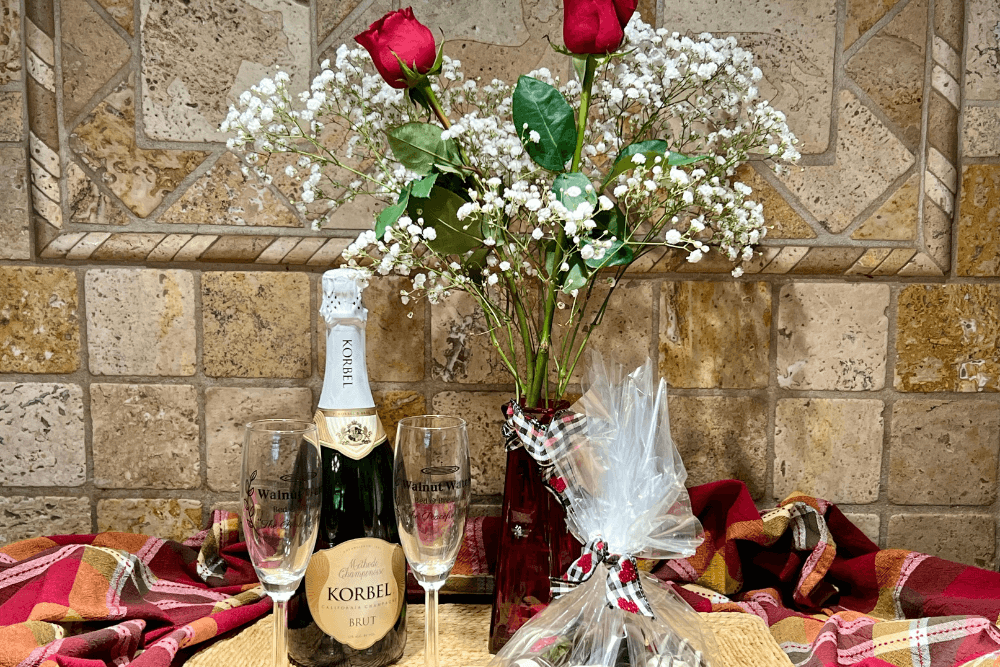 Vase of red roses with baby's breath, bottle of champagne with two flutes and cellophane-wrapped chocolate covered strawberries.