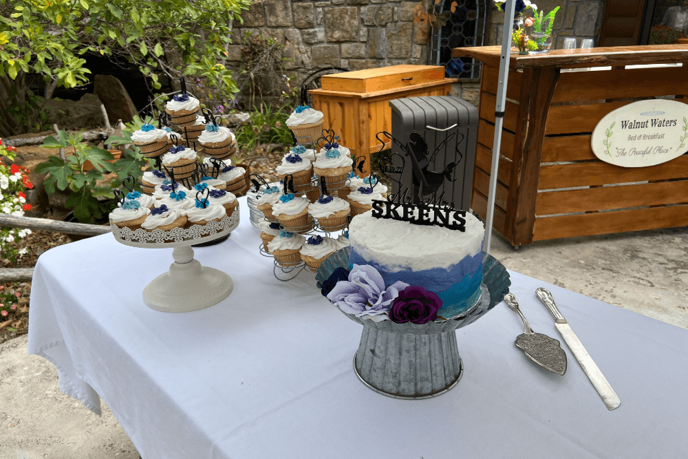 Table outdoors with white tablecloth with 3 cake stands with cupcakes and cakes with a silver serving knife and cake server.