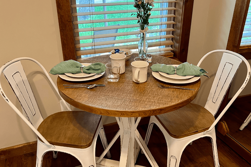 Round wood table with two white chairs, set for a breakfast for two, with white plates and cups, green napkins and a vase of flowers