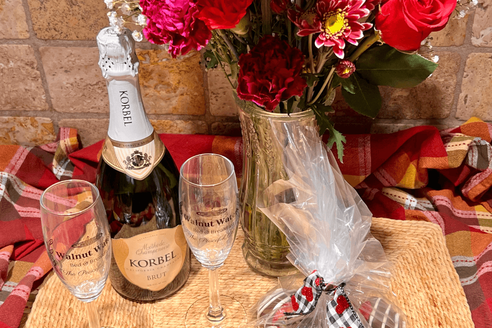 Table with bottle of champagne, two flutes, vase of yellow flowers and wrapped plate of desserts