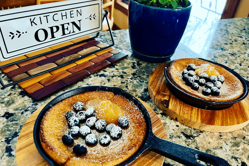 Two cast-iron skillets filled with a breakfast dish topped with blueberries and powdered sugar on a kitchen counter