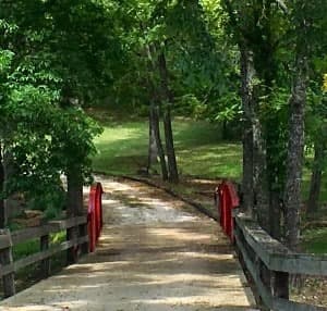 A winding path leads over a small wooden bridge with red railings, surrounded by lush green trees.