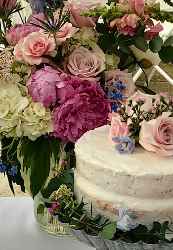 Wedding cake topped with pink flowers and a vase of multi colored flowers behind it