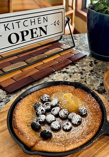 Cast Iron frying pan with pancake topped with blueberries . A sign that says Kitchen Open and a plant in a blue pot