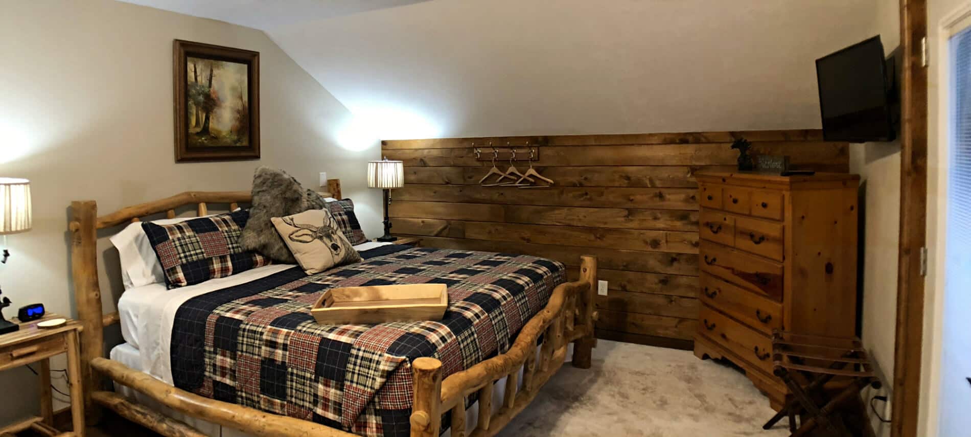 Walnut guest room with sloped ceiling, short plank wall, rustic bed with plaid bedding, and TV
